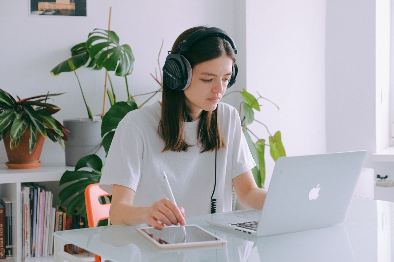 Young woman in home office using a laptop and drawing tablet, wearing headphones, illustrating remote work.